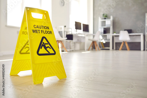 Close up shot of plastic standing sign that reads Caution Wet Floor and has figure of man who slips and falls office interior in blurred background Fotobehang