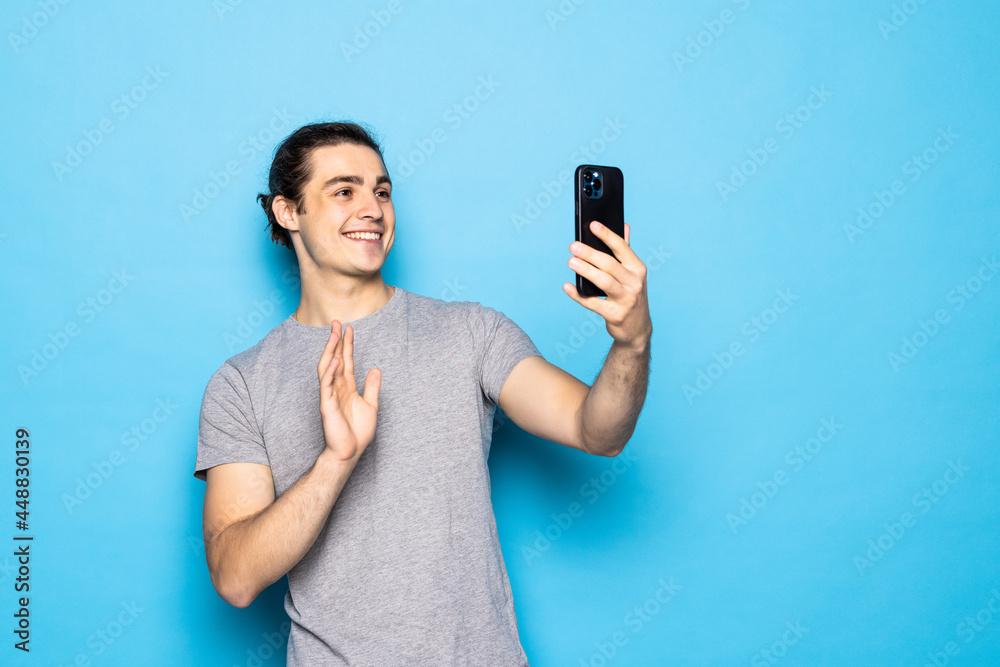 Happy young man video calling, talking online with mobile phone, saying hello to smartphone camera and waving hand friendly, standing over light blue background Happy young man video calling, talking online with mobile phone, saying hello to smartphone camera and waving hand friendly, standing over light blue background