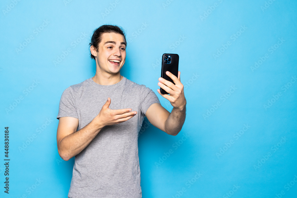 Happy young man video calling, talking online with mobile phone, saying hello to smartphone camera and waving hand friendly, standing over light blue background Happy young man video calling, talking online with mobile phone, saying hello to smartphone camera and waving hand friendly, standing over light blue background