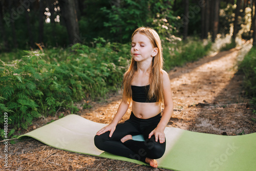 A beautiful blonde preschool girl, a child athlete professional in a black suit sits on a green rug in a lotus position, crossing her legs, and meditates, relaxes, training yoga exercises.