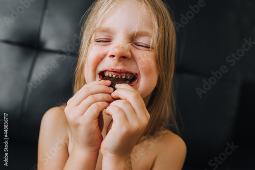 Close-up portrait of a funny little girl  a child  who is smeared in black  milk chocolate and emotionally bites it. Photography  concept.