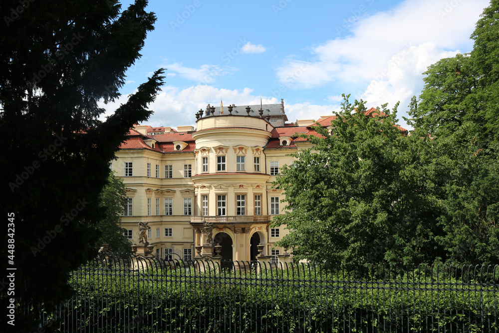 Lobkovicz Palace in Prague illuminated by the bright sun among the greenery of trees against the background of an azure sky and light clouds