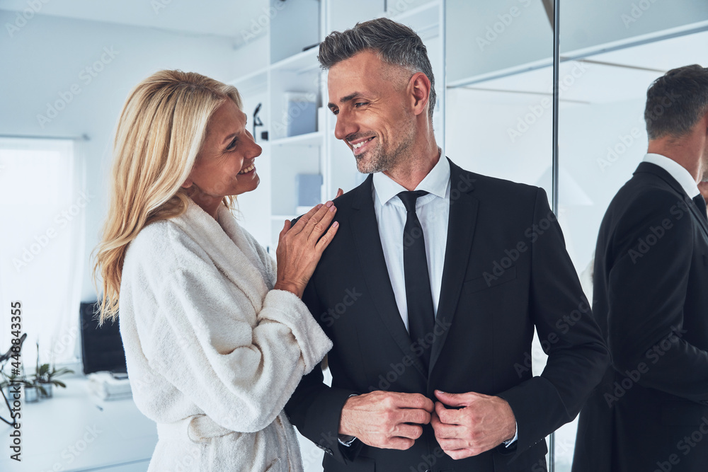 Beautiful mature woman adjusting suit of her husband while both standing in front of the mirror at home