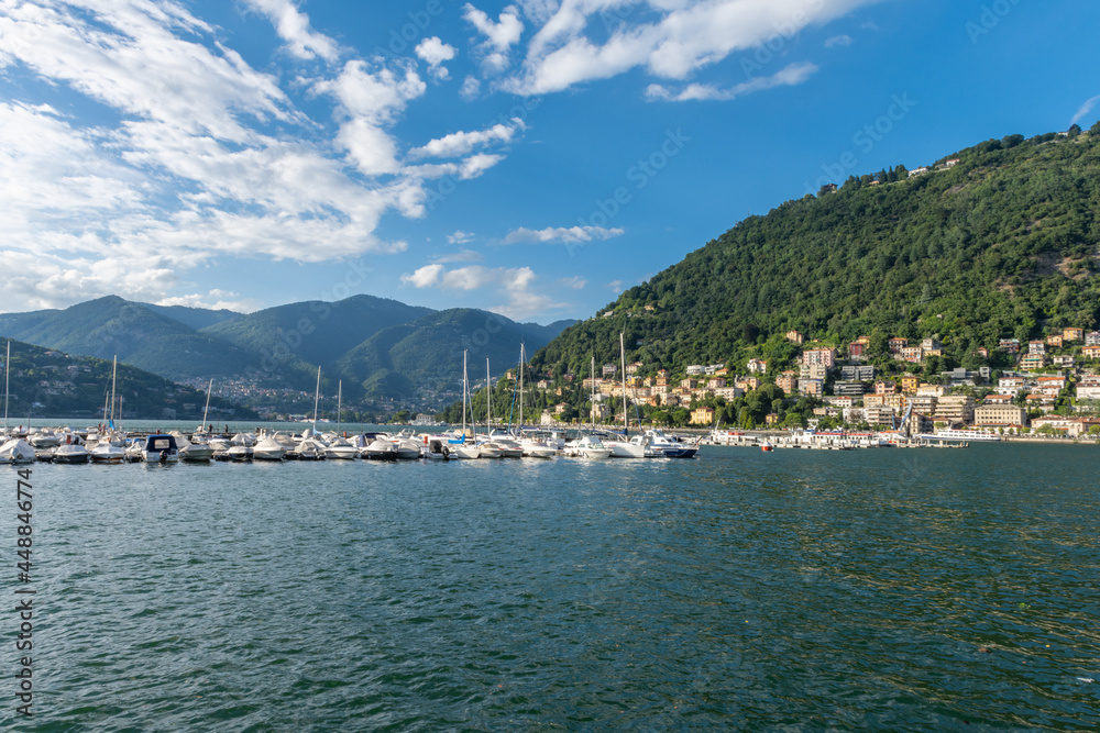 View over the beautiful, gorgeous lake Como and the marina seen from the town of Como. It is a beautiful sunny summer day, with blue sky and a few clouds. There are many boats and dinghy in the marina