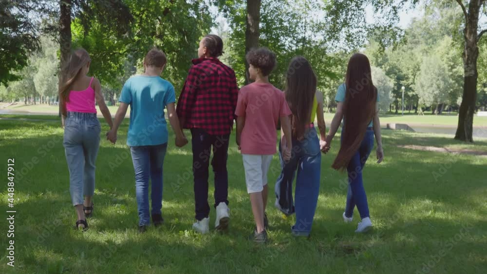 Follow shot of diverse teen international friends hold hands walking in park Follow shot of diverse teen international friends hold hands walking in park