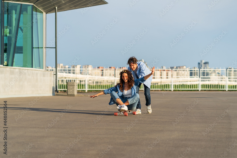 Obraz premium Young caucasian couple enjoy skating together on street. Hipster man pushing back of woman sitting on longboard. Cheerful lovers or friends have fun outdoors over city skyline. Urban lifestyle concept