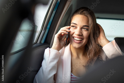 Portrait of a beautiful business woman using a smart phone and smiling while sitting on the back seat of the car. Young woman with smartphone on the back seat of a car