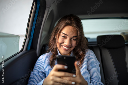 Smiling casual woman sitting in back seat using mobile phone. Cheerful young woman reading messages in smartphone while sitting in a taxi. Attractive girl wearing blue shirt in car using cellphone.