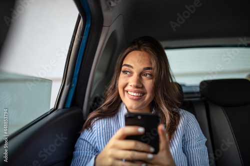Smiling casual woman sitting in back seat using mobile phone. Cheerful young woman reading messages in smartphone while sitting in a taxi. Attractive girl wearing blue shirt in car using cellphone. © Dragana Gordic