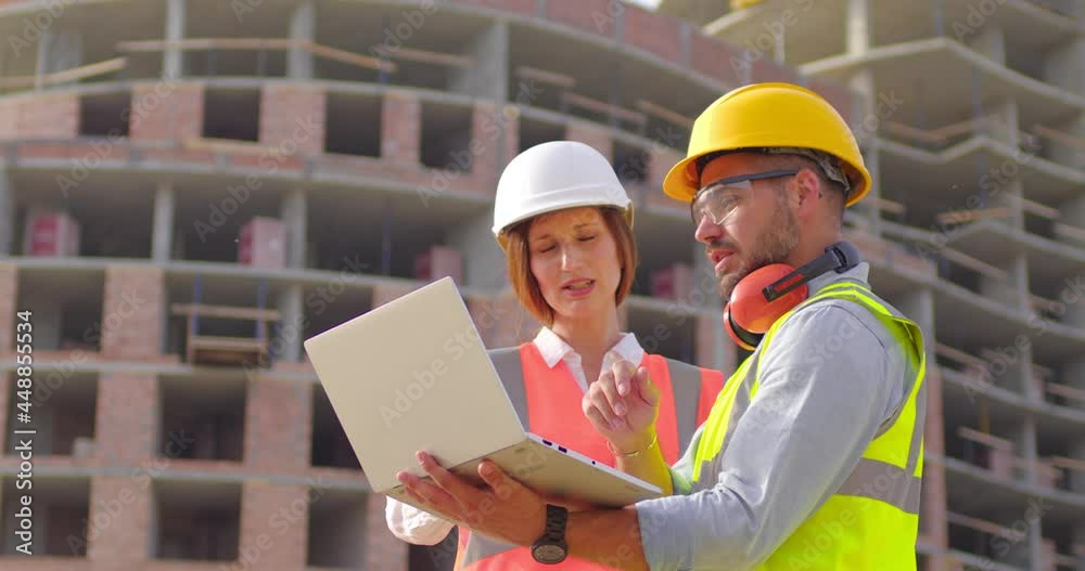 Young foreman and female architect standing in front of construction site and look at the lap top, discussing their job. The concept of new technologies in construction. Front view. Close up. Young foreman and female architect standing in front of construction site and look at the lap top, discussing their job. The concept of new technologies in construction. Front view. Close up.