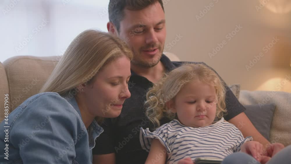 Family sitting on sofa at home as young daughter plays game on mobile phone - shot in slow motion Family sitting on sofa at home as young daughter plays game on mobile phone - shot in slow motion