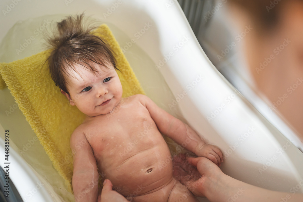 Naklejka premium Small caucasian baby taking a bath at home - unknown woman mother holding her small child baby in plastic bath tub in room at home parenting and hygiene concept real people