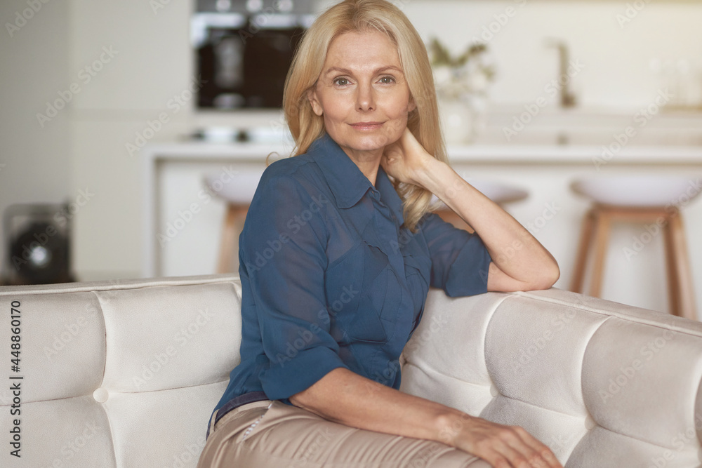 Attractive mature woman with pleasant smile looking at camera while relaxing, sitting on sofa in her modern apartment Attractive mature woman with pleasant smile looking at camera while relaxing, sitting on sofa in her modern apartment