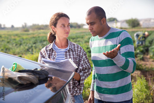 Focused couple of farmers standing outdoors near car on background with vegetable plantation, discussing documents.. © JackF