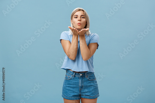 Studio shot of attractive and romantic european blonde girlfriend in denim shorts folding lips sending wind kiss with palms near mouth gazing tender and cute at camera over blue background photo