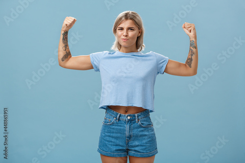 Strong and powerful good-looking sportswomen with tattoos in t-shirt and shorts raising arms showing big muscles and biceps smiling proudly while bragging with physical strength over blue wall