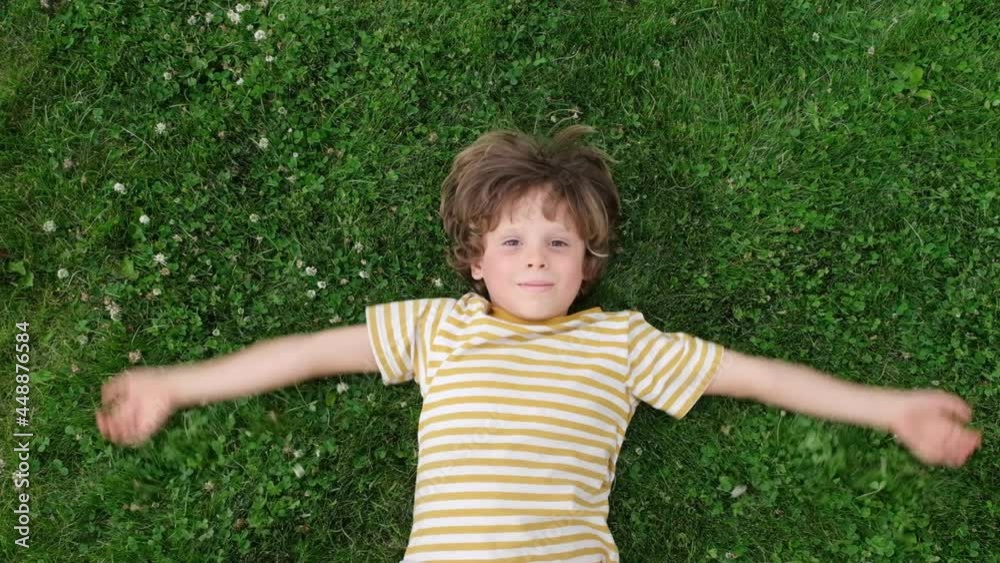 Cute funny child boy is lying on green grass, top view. Happy boy making an angel like in the snow. kid looking at camera and smiling. View from above. Slow motion. Cute funny child boy is lying on green grass, top view. Happy boy making an angel like in the snow. kid looking at camera and smiling. View from above. Slow motion.