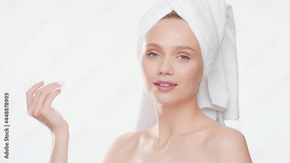 Gorgeous European young slim woman wrapped in towel touches her cheek with cotton pad after taking a bath, turns her head to the camera and smiles wide for it on white background | Anti-aging concept