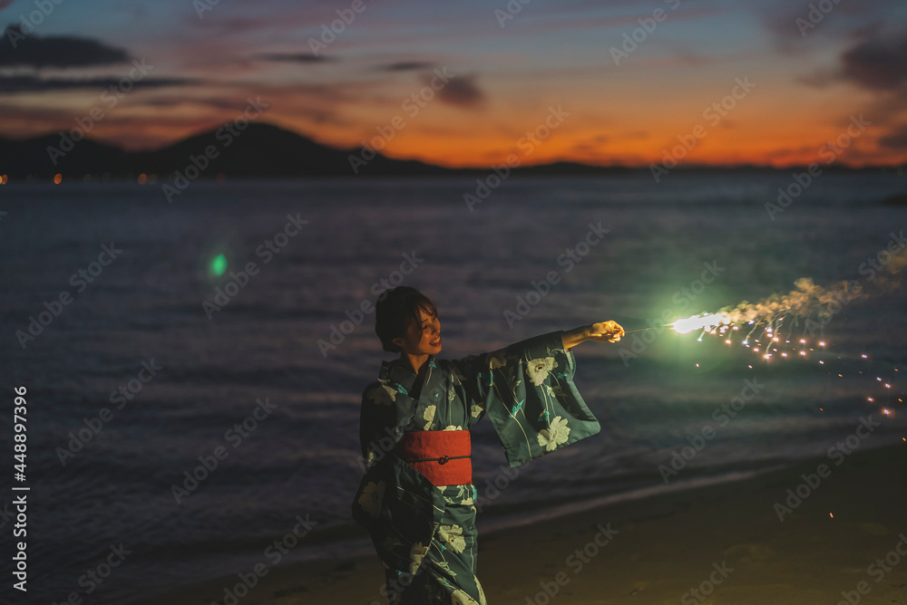 Obraz premium 夕暮れの海で花火をする浴衣の女性