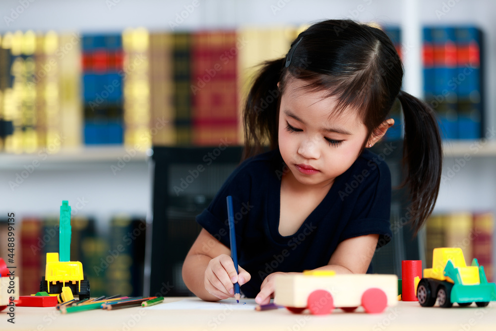 Small pretty pigtails hair preschooler kindergarten happy girl sit on chair drawing cartoon with colored pencils on paper on table full of plastic truck toys in living room at home in front bookshelf Small pretty pigtails hair preschooler kindergarten happy girl sit on chair drawing cartoon with colored pencils on paper on table full of plastic truck toys in living room at home in front bookshelf