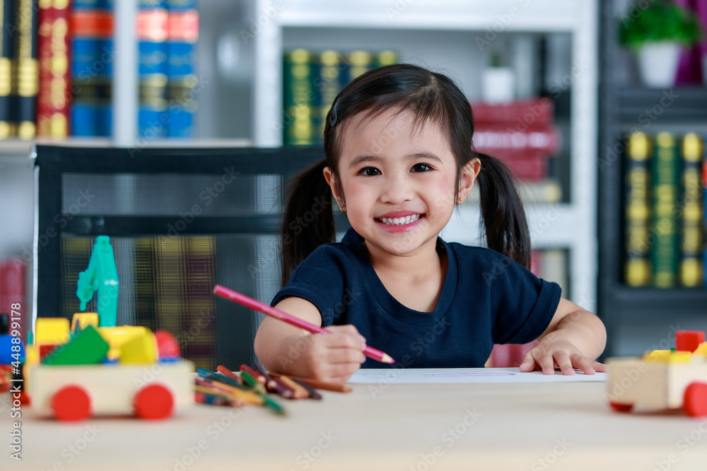 Small pretty pigtails hair preschooler kindergarten happy girl sit on chair drawing with colored pencils on paper on table full of plastic truck toys in living room at home in front bookshelf Small pretty pigtails hair preschooler kindergarten happy girl sit on chair drawing with colored pencils on paper on table full of plastic truck toys in living room at home in front bookshelf