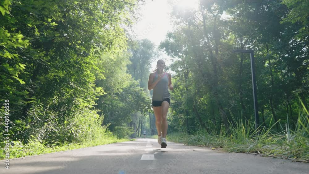 Slow motion sporty woman jogging on asphalt road among green trees. Tracking shot female athlete training in morning sunshine. Concept of fitness