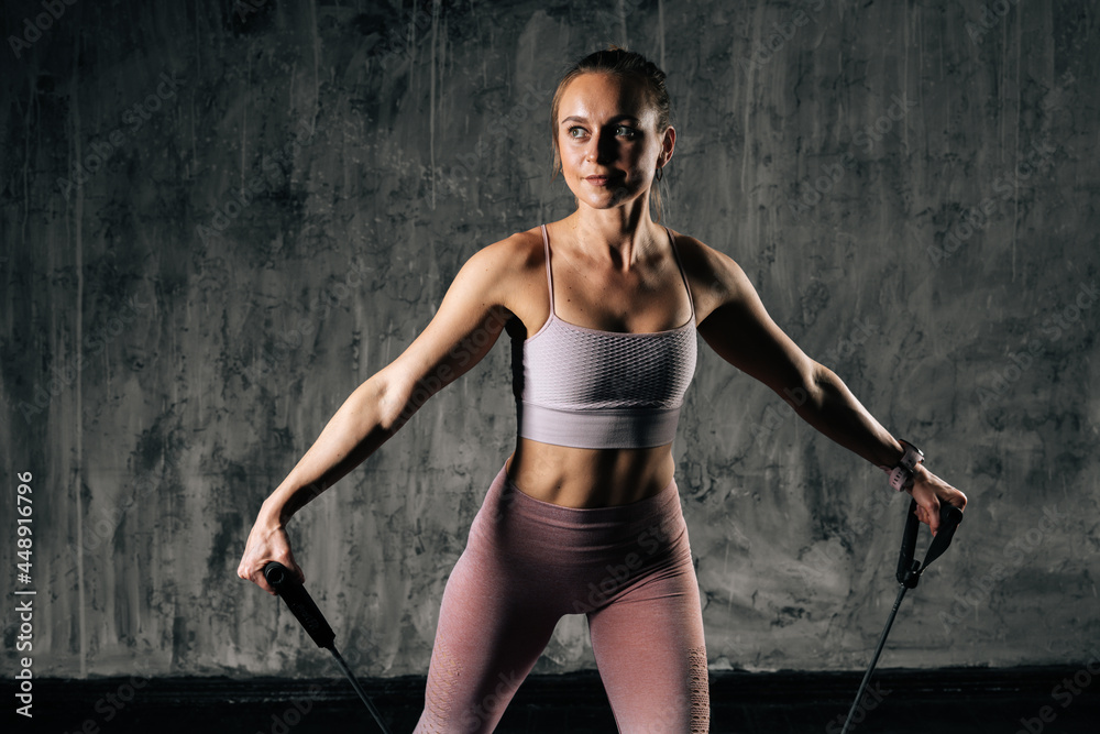 Muscular young athletic woman with perfect beautiful body wearing sportswear exercising with resistance band standing on mat. Caucasian fitness female training with stretching expander in studio. Muscular young athletic woman with perfect beautiful body wearing sportswear exercising with resistance band standing on mat. Caucasian fitness female training with stretching expander in studio.