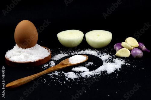 Salt on the wooden spoon and standing egg on the salt plate with some onions in black background