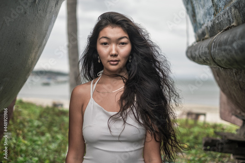 Portrait of beautiful young mongol woman in white dress standing between two old wooden ships. Brunette long curly hair. Looking at camera with copy space. Romantic portrait.