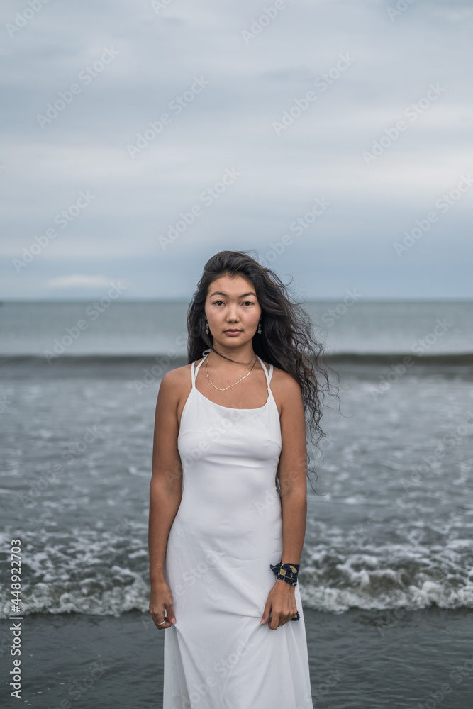 Portrait of charming young mongol woman in bright grey dress standing on the beach. Black long curly hair. Looking at camera with copy space. Romantic photo. Portrait of charming young mongol woman in bright grey dress standing on the beach. Black long curly hair. Looking at camera with copy space. Romantic photo.