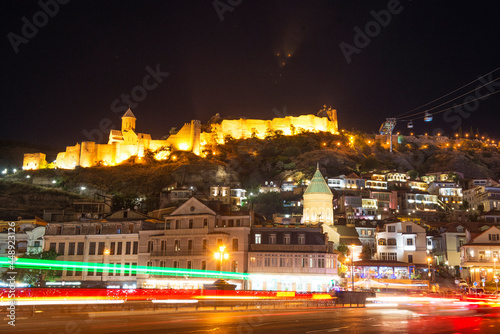 Night city of Tbilisi. In the background is a cascade of buildings with architecture typical of the region. Narikala Fortress is illuminated by the initial lighting. In the foreground are long exposur