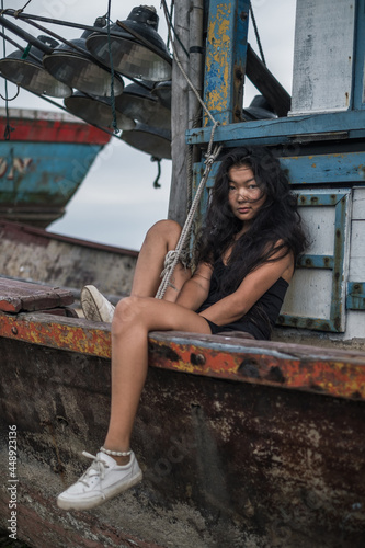 Charming young mongol woman in black t-shirt and white sneakers sitting on the old wooden ship and holding her legs. Brunette long curly hair. Romantic photo.