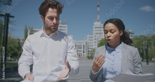 Diverse business colleagues discussing papers and quarrelling, having conflict about contract details, walking outdoors photo