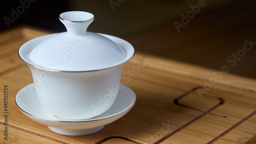 Gaiwan on chaban selective focus. Tea ceremony gong fu cha. Chinese white pottery Dehua white Chinese porcelain gaiwan bowl tea cup and lid plate with gold border on bamboo teaboard tea tray chaban photo