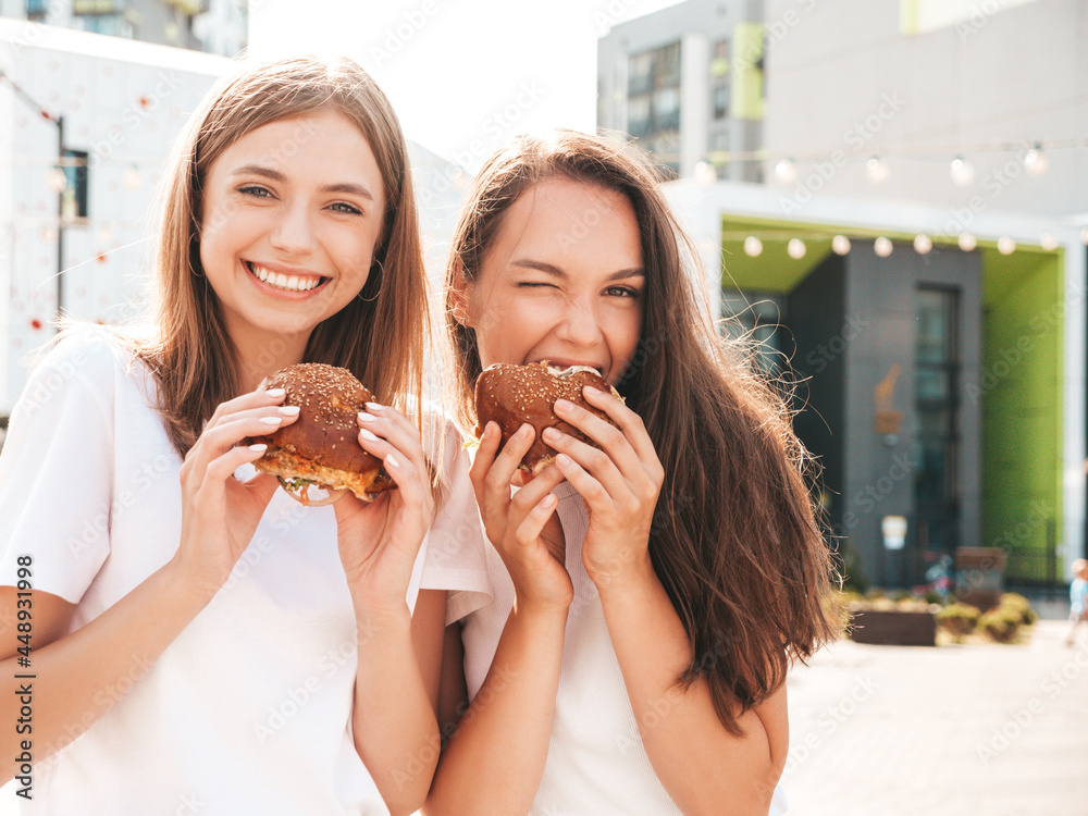 Two young beautiful smiling hipster female in trendy summer same clothes. Sexy carefree women posing in the street.Positive models having fun in sunglasses.Holding juicy burger and eating hamburger Two young beautiful smiling hipster female in trendy summer same clothes. Sexy carefree women posing in the street.Positive models having fun in sunglasses.Holding juicy burger and eating hamburger