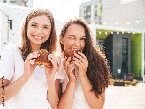 Two young beautiful smiling hipster female in trendy summer same clothes. Sexy carefree women posing in the street.Positive models having fun in sunglasses.Holding juicy burger and eating hamburger