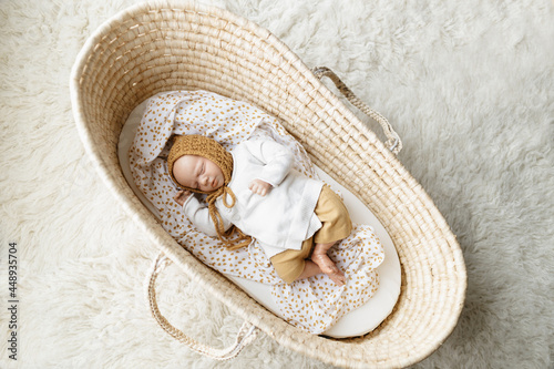 Fotografie A reborn newborn baby doll toy dressed in a mustard colored bonnet hat and a white cardigan lying in a cream moses basket bassinet