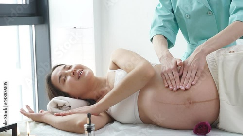 Professional massage therapist massages arm and shoulders of young pregnant woman lying on massage table, side view. Beautiful gravid female relaxing massage. Concept of body care.