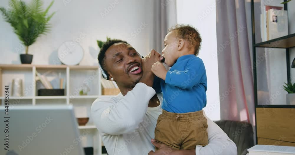 Distant work and child care concept where handsome smiling caring skilled black-skinned dad talking on video chat with colleagues when his small kid playing with microphone attached to father's Distant work and child care concept where handsome smiling caring skilled black-skinned dad talking on video chat with colleagues when his small kid playing with microphone attached to father's
