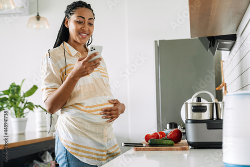 Black pregnant woman smiling and using mobile phone in kitchen