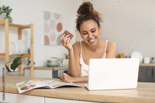 Young African-American woman with chocolate using laptop in kitchen