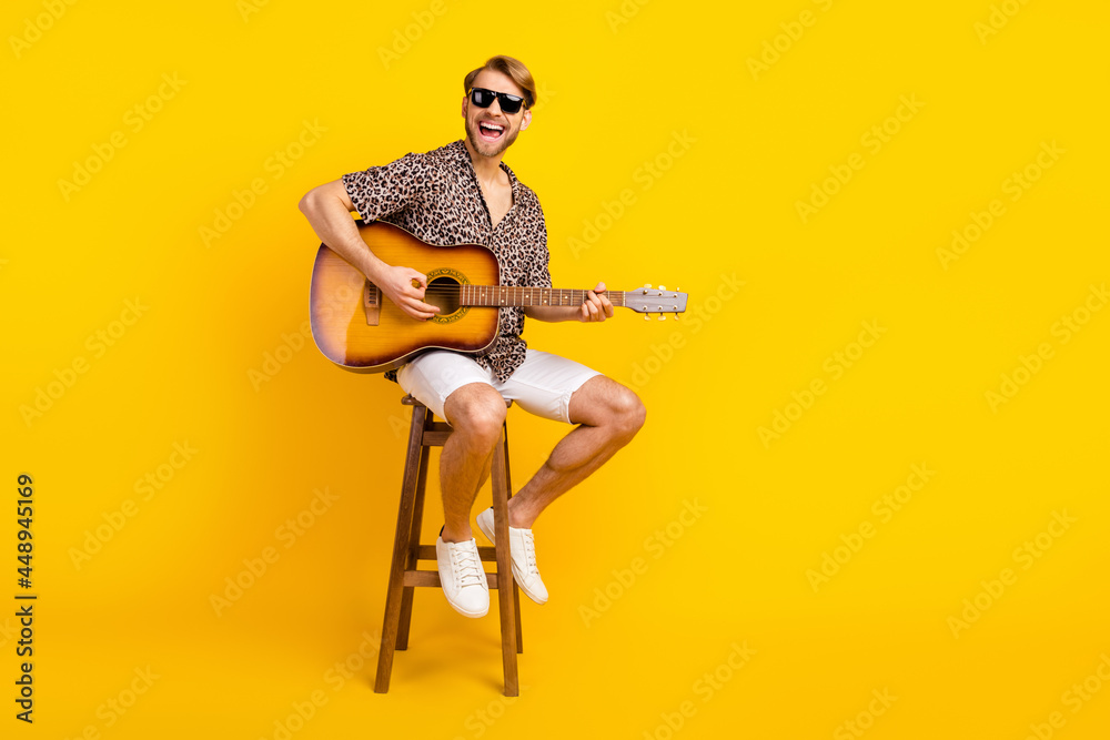 Photo of sweet funny young gentleman dressed print shirt dark glasses sitting bar chair playing guitar singing isolated yellow color background Photo of sweet funny young gentleman dressed print shirt dark glasses sitting bar chair playing guitar singing isolated yellow color background