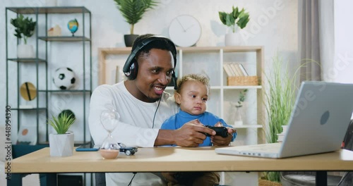 Child care concept where attractive happy carefree loving young black-skinned man having fun together with his 2-aged son in videogames on laptop at home photo