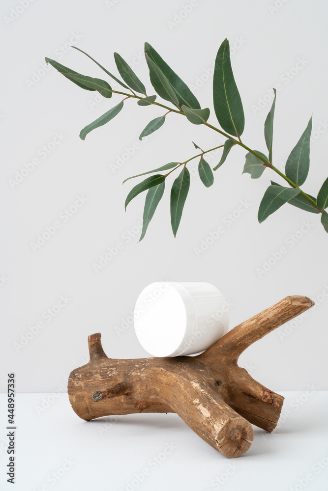 Naklejka premium White container for branding on wooden stick podium and eucalyptus branch on grey background. Piece of wood, green leaves and cosmetic mockup. Rough natural snag and beauty product advertising. 