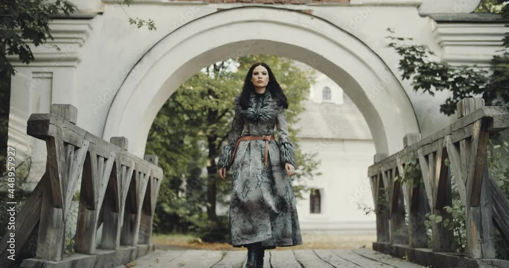 View of charming woman going under ancient arch in a castle, passing wooden bridge. Leather coat with fur, gothic or scandinavian style. summer nature background