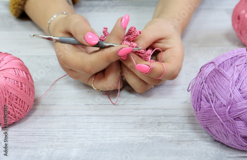 The concept of crocheting products.Crochet hook in women's hands with a pink manicure among multi-colored skeins, gray background, side view