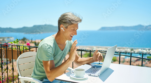 Joyful man wearing headphones communicating with friends via video computer call during sea resort summer vacation