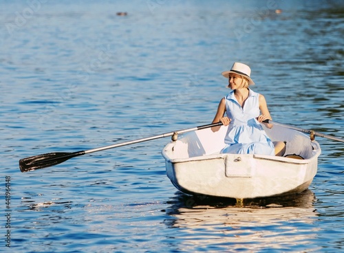 An elegant blonde woman in a hat sits on a boat and enjoys the tranquil lake during her summer vacation.