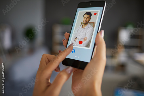 Female finger pushing chat button on smartphone using dating app to find love and online connection. Focus on handsome man profile photo on screen. Modern lifestyle relationship, remote relation