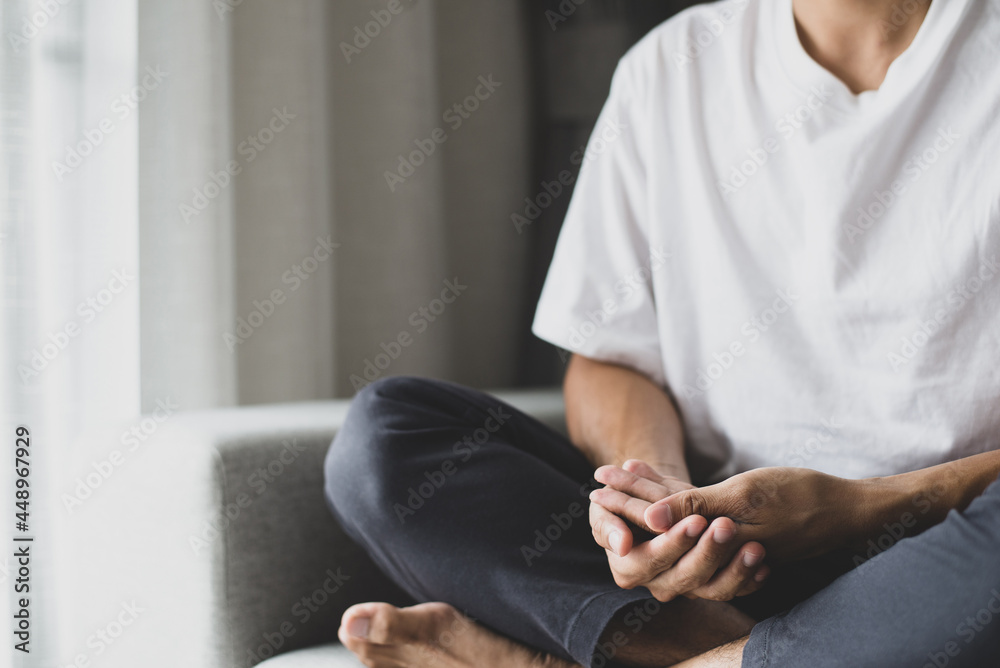 Young man in a white t-shirt and grey pants sat on the sofa practicing meditation, sitting in a lotus pose alone in the morning at home. During the quarantine due to the spread of the coronavirus.